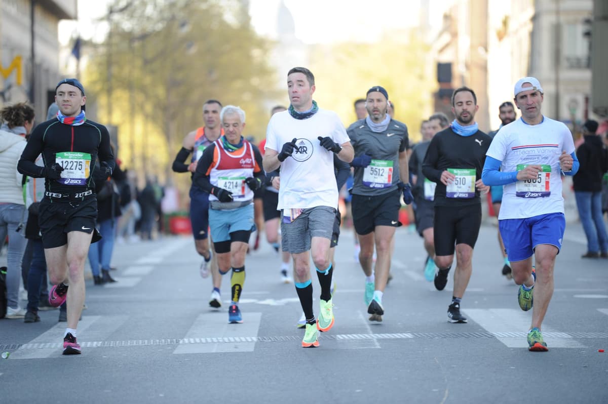 Group of marathon runners with race bibs running down a city street during Paris Marathon 2022, with buildings and trees in background
