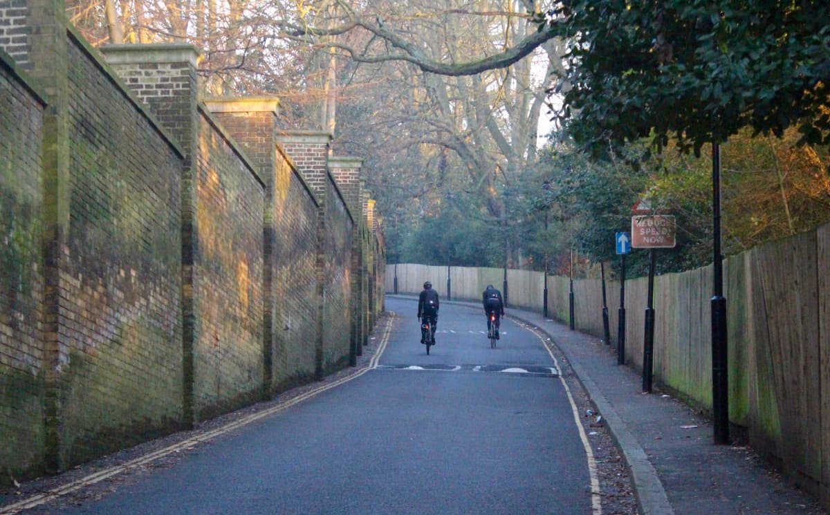 Two cyclists riding up a tree-lined road with brick walls on both sides, autumn foliage overhead on what appears to be Swains Lane in London