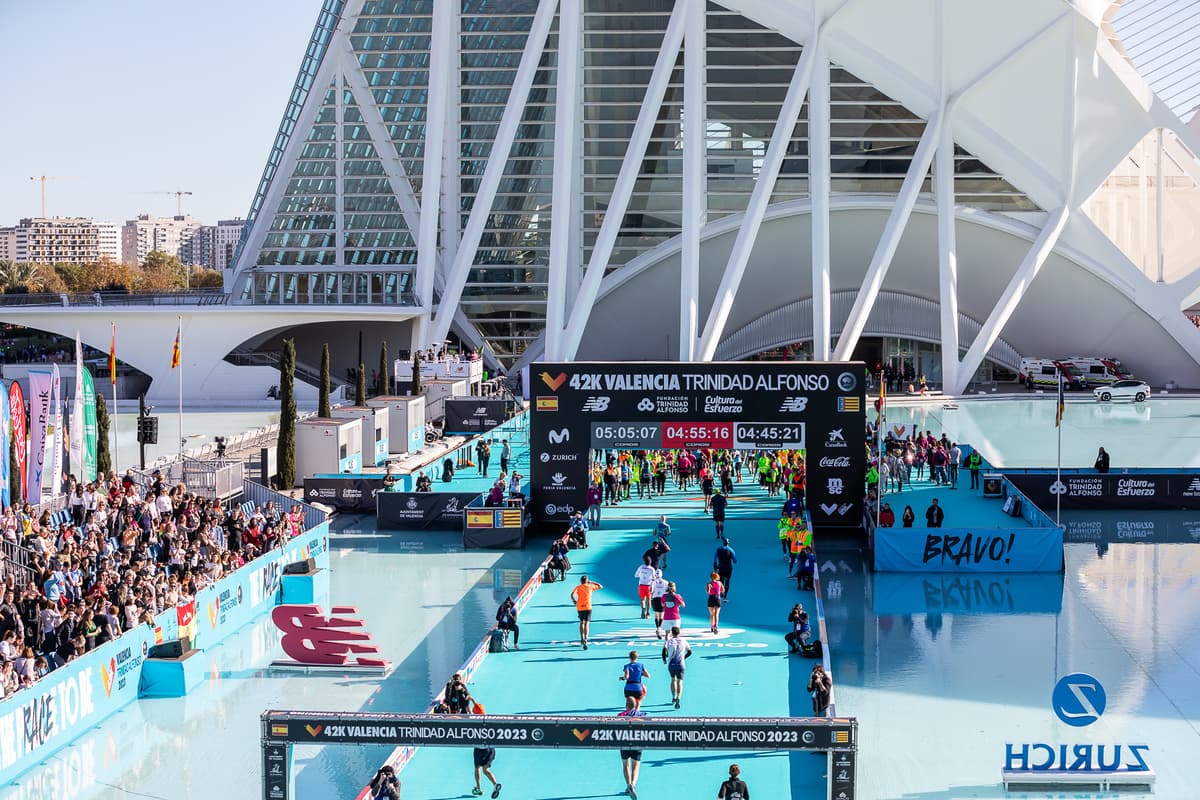 Marathon finish line at Valencia Trinidad Alfonso 42K with runners crossing under timing displays showing race times, surrounded by cheering crowds and modern architecture