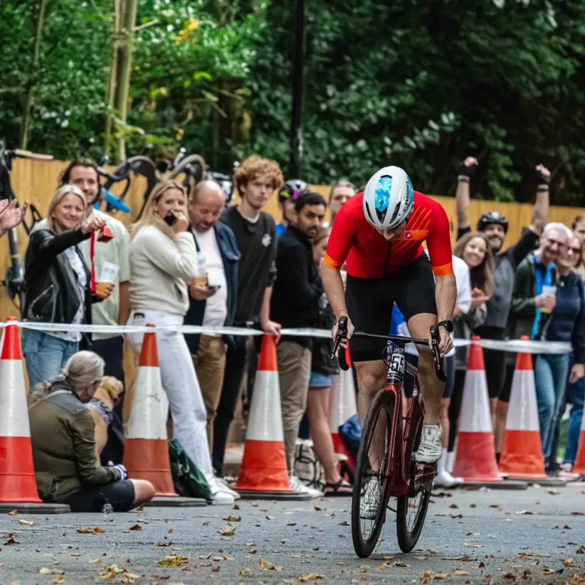 Cyclist in red jersey and aerodynamic helmet racing on time trial bike past cheering spectators lined behind orange traffic cones